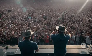 Two men in hats on a stage face a massive, cheering crowd at an outdoor concert.