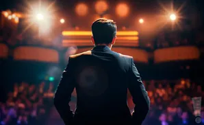 A person in a dark suit stands facing a brightly lit audience on a stage.
