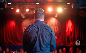 Man in a dark shirt stands on stage facing a red curtain and audience, lit by stage lights.