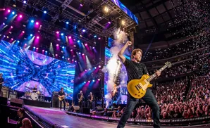 A male guitarist on stage rocks out as confetti rains down on a cheering crowd at a concert.