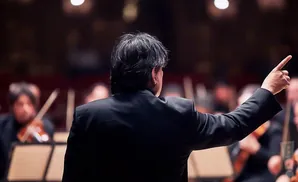 Conductor in a black suit with his back to the camera, pointing with his right hand to the right.