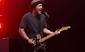 A man wearing a hat and dark t-shirt plays a guitar and sings into a microphone on a red-lit stage.