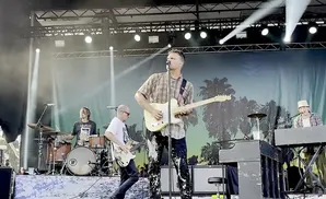 A band performs on stage with a palm tree backdrop. The lead singer plays a light-colored electric guitar.