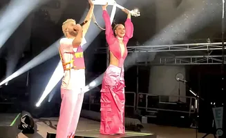 A man and woman on stage, the man holds a microphone and the woman holds a guitar above her head.