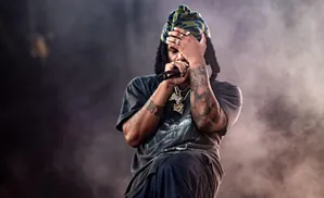 A rapper with a beanie, jewelry, and tattoos holds a microphone and covers their face.