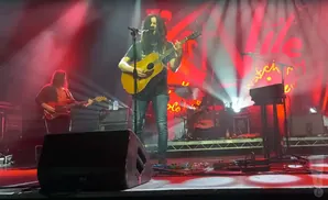 A guitarist with long hair plays an acoustic guitar on stage with red lighting. Another musician plays bass to the left.