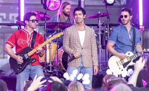 Three men on stage with instruments, one in a red baseball jersey, one in a plaid jacket, and one in a denim shirt.