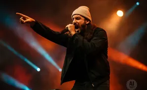 A man with long hair and a beanie sings into a microphone on a stage with colorful lights.
