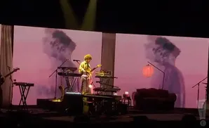 A musician with curly hair plays a guitar on stage in front of a large screen displaying abstract, hazy images.