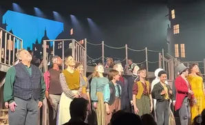 A group of performers in historical costumes stand on a stage, looking up at the audience.