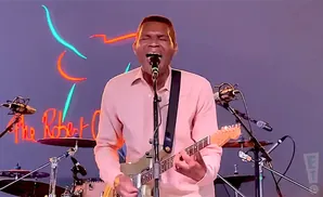 A man in a pink shirt sings and plays a guitar in front of a neon sign that says 