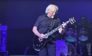 Musician with white hair playing a black electric guitar on stage with a drum set in the background.