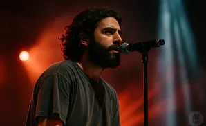 A man with dark, curly hair and a beard sings into a microphone on stage with red and white stage lights.