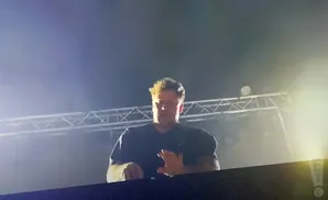 A DJ with light-colored hair and a black t-shirt stands behind a dark console, silhouetted by stage lights.
