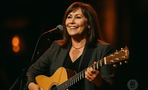 A woman with brown hair smiles while playing an acoustic guitar and singing into a microphone.