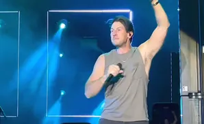 A male singer with arms raised, holding a microphone and wearing a grey tank top on a blue-lit stage.