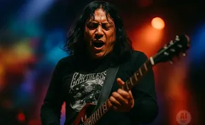 A man with long dark hair passionately sings while playing a guitar on a colorful stage.