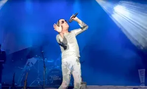 A singer in a silver outfit and sunglasses performs on a blue-lit stage, holding a microphone.
