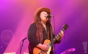 A person in a cowboy hat sings into a microphone while playing a guitar on a purple-lit stage.