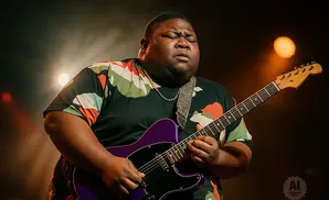 A man plays a purple electric guitar, eyes closed, on a dimly lit stage.