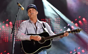 A man in a striped shirt and baseball cap plays an acoustic guitar on stage with red lights in the background.