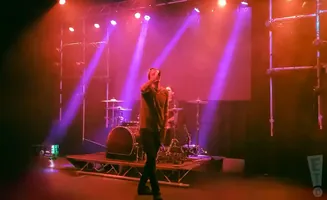 A male singer performs on stage with a drum set in the background, illuminated by vibrant pink and red stage lights.