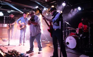 A band plays live music on a stage with colorful stage lighting and smoke.