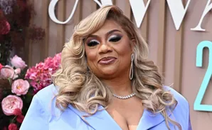 A woman with blonde curly hair, heavy makeup, and a blue dress smiles.