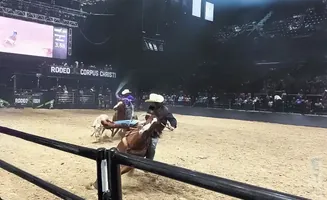 Rodeo riders on horseback in an arena, with a large screen displaying scores and crowd seating in the background.