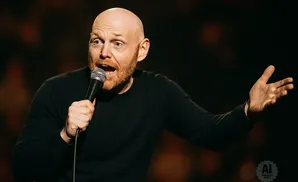 Bill Burr, a bald comedian with a red beard, holds a microphone and gestures with his left hand on stage.