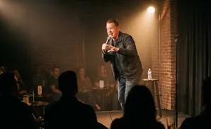 A smiling man in a black suit and tie holds a vintage microphone, standing against a blue background.