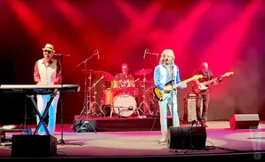 A band performs on a stage with red lighting: a keyboardist, drummer, guitarist, and bassist.