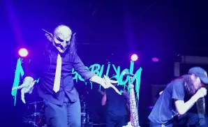 A performer in a goblin mask and tie sings into a microphone on a dimly lit stage.