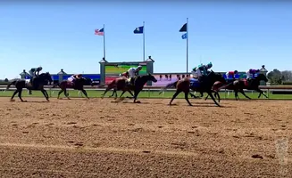 Horses and jockeys race on a dirt track under a clear blue sky, with flags and a large screen in the background.
