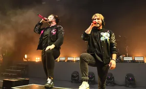 Two male singers perform on stage in black jackets, with spotlights and smoke in the background.