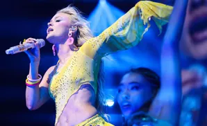 A singer in a sparkly yellow outfit sings into a bejeweled microphone, with a dancer blurred in the background.