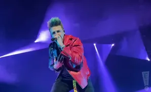 A man with spiky green hair and a red jacket sings into a microphone on a purple-lit stage.