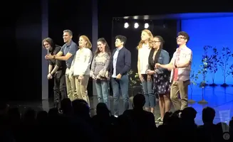 A group of actors stand on a stage, smiling at an audience.