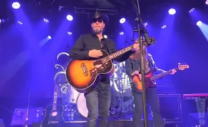 Man in cowboy hat plays acoustic guitar on stage with blue lights.
