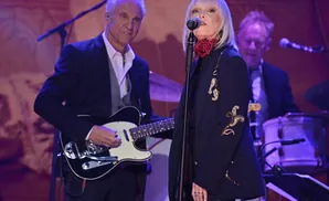 Singer with silver hair performs on stage, wearing a dark jacket with gold embroidery, while a guitarist plays beside her.