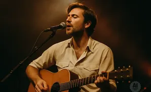 Man playing an acoustic guitar and singing into a microphone on a dimly lit stage.