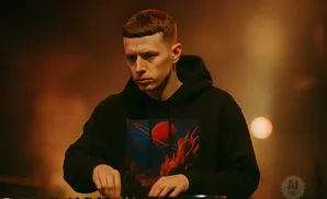 A DJ with a bowl cut, wearing a black hoodie with a fiery planet design, focuses on his equipment.