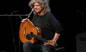 A man with wild, curly grey hair plays an acoustic guitar on stage.