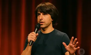 A man with dark hair speaks into a microphone, holding his left hand up.