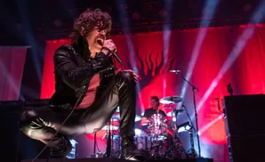 A male rock singer with curly hair and a mustache crouches and sings into a microphone on a stage with red lighting.