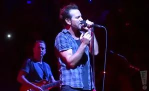 A man in a blue plaid shirt sings into a microphone on stage, with another man playing guitar behind him.