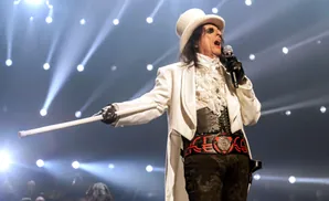 Alice Cooper in a white top hat and tails performs on stage with a microphone and a white cane.