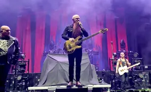 A bald bassist with a long beard plays a gold bass guitar on stage, with two other men standing beside him.