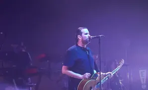A man sings into a microphone and plays an acoustic guitar on a stage with purple lighting.