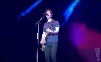 A male guitarist plays his electric guitar on stage under blue and pink spotlights.